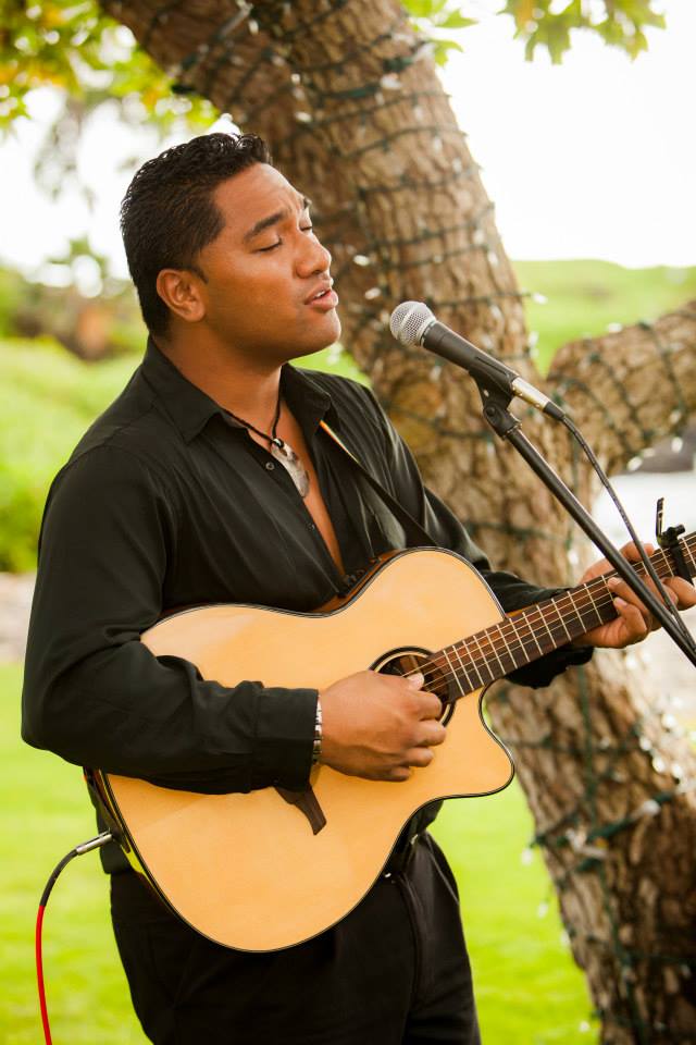Kaniala Masoe singing into a microphone while playing an acoustic guitar beside a palm trunk wrapped in string lights