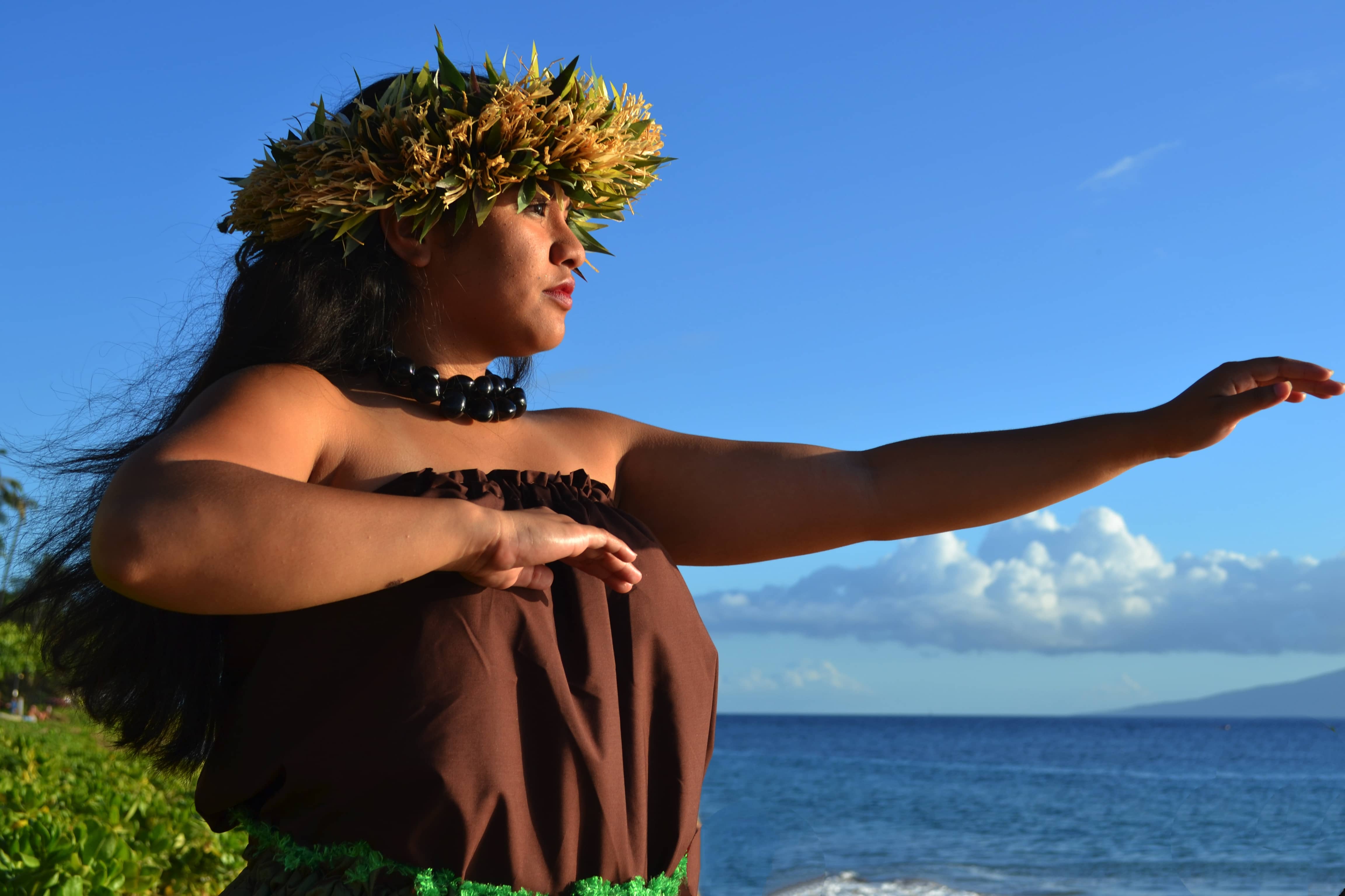 Melody Masoe dancing hula in a haku lei of green ti leaves and a kukui nut lei, one arm extended toward the ocean under a clear sky